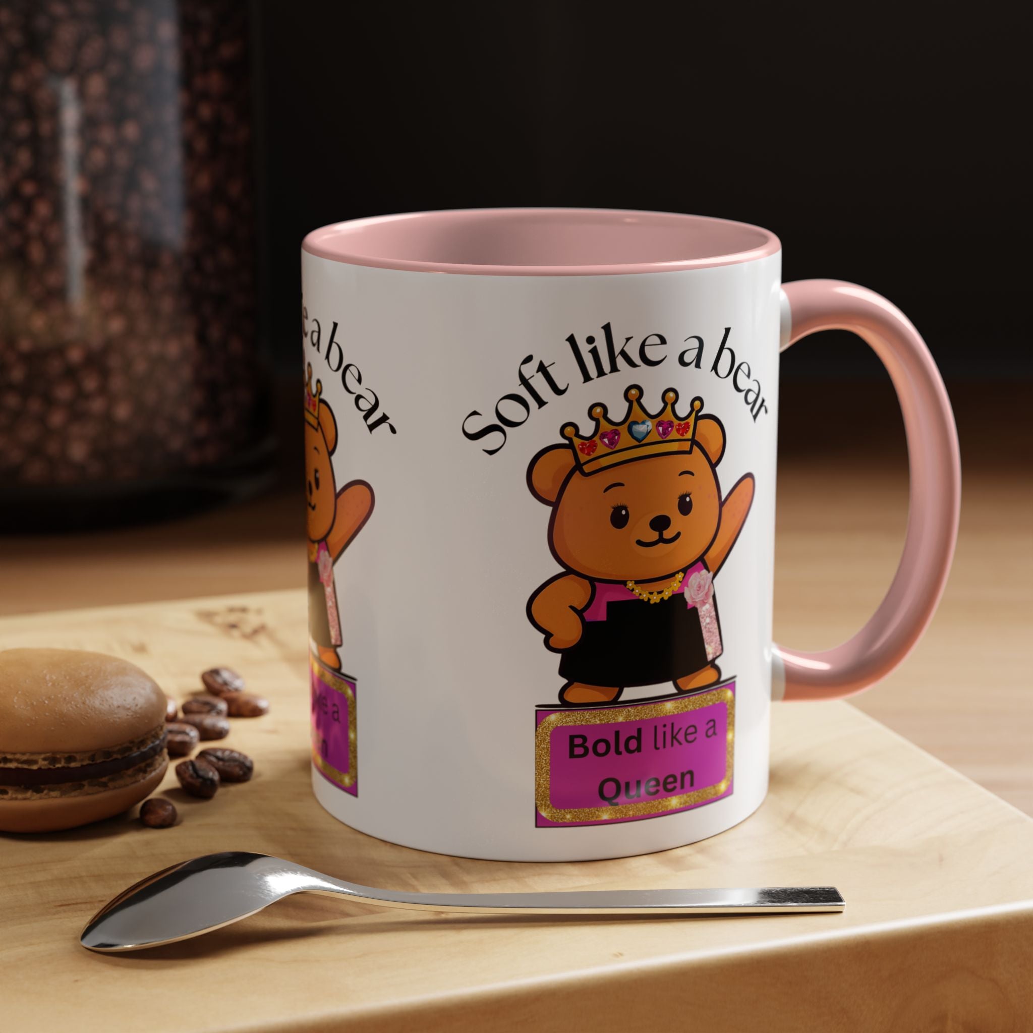 White mug with pink interior and handle, featuring a cartoon bear design and text, on a wooden surface with coffee beans and a spoon.